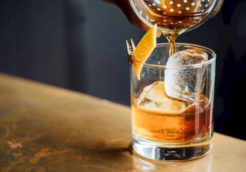 A glass of whiskey on the rocks being poured with an orange twist garnish, tipped into a lowball glass.