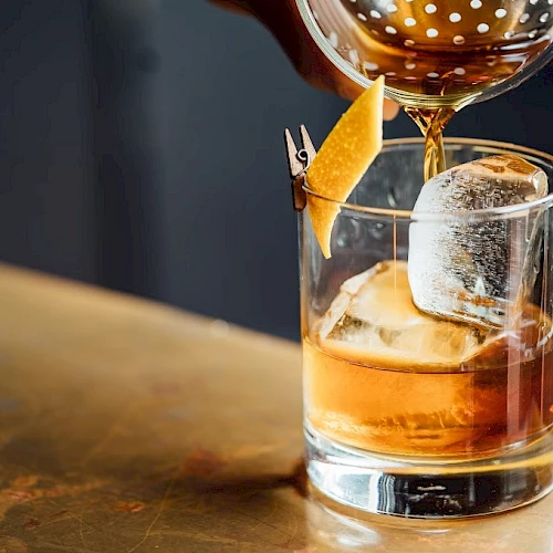 A glass of whiskey on the rocks being poured with an orange twist garnish, tipped into a lowball glass.