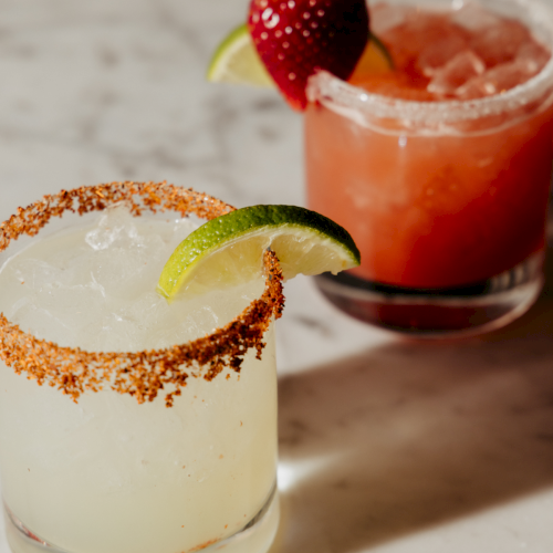 Two cocktails with sugar rims and fruit garnish (one lime, one strawberry) on a marble surface. Top it at 140 characters, always ending the sentence.