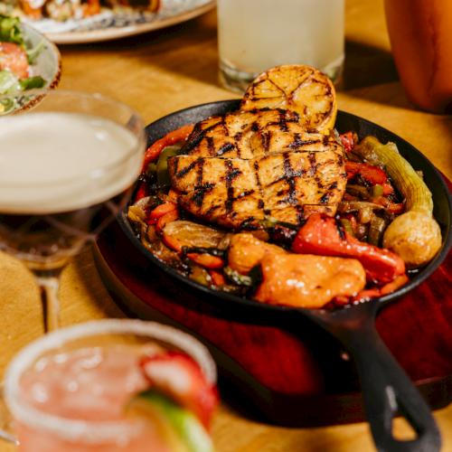 A spread of grilled meats and seafood with sides, plus drinks (a citrus cocktail, a pink drink, and a light-colored beverage) on a wooden table.