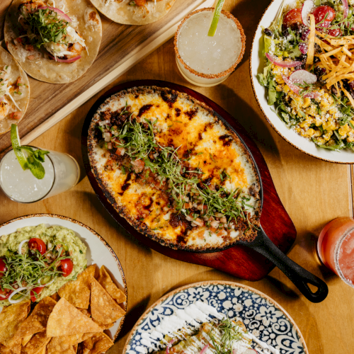 A spread of colorful dishes: a skillet with pizza or calzone, bowls of dips, fresh salads, and toppings, all on a warm wooden table.