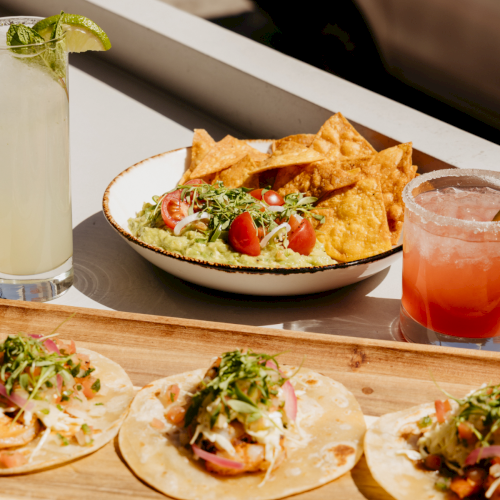A colorful plate of tacos with toppings, a lime wedge, and fresh herbs, plus a small drink on a wooden table, Mexico-inspired feast.