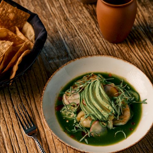 A small bowl of green dip (pesto or guacamole) with tortilla chips and walnuts on the side, in a rustic setting.