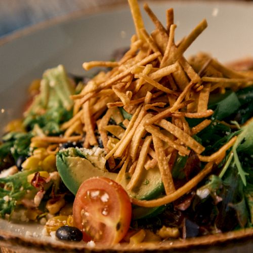 A vibrant salad with greens, avocado, cherry tomato halves, black beans, and crunchy fried tortilla strips on top, in a rustic bowl.