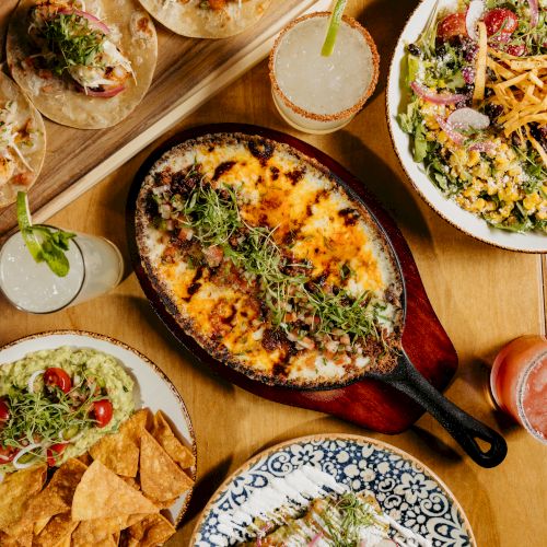 A spread of tacos, a sizzling skillet of cheesy skillet dip, a fresh salad, tortilla chips with salsa, and drinks on a wooden table.