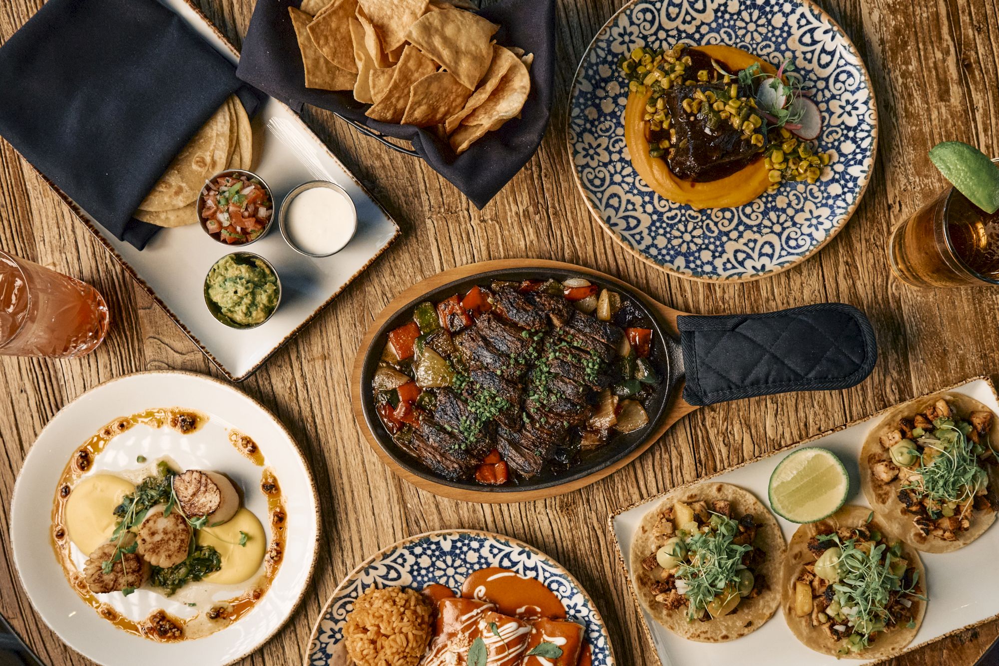 A spread of sizzling, tapas-style dishes and dips on a wooden table, ready for sharing, with plates, bowls, and sauces all around.