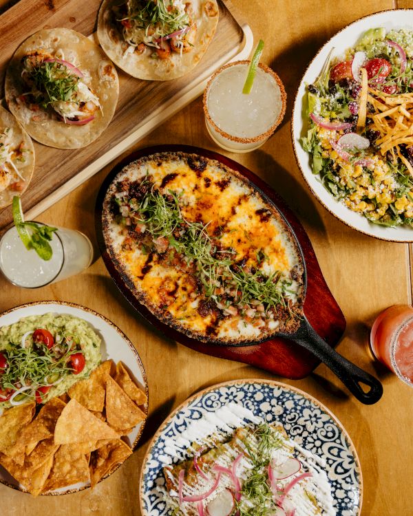 A spread of tacos, a sizzling skillet of cheesy skillet dip, a fresh salad, tortilla chips with salsa, and drinks on a wooden table.