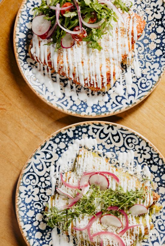 Two plates of plated grilled fish with ribbons of sauce, microgreens, and pickled onions on a decorative patterned plate.