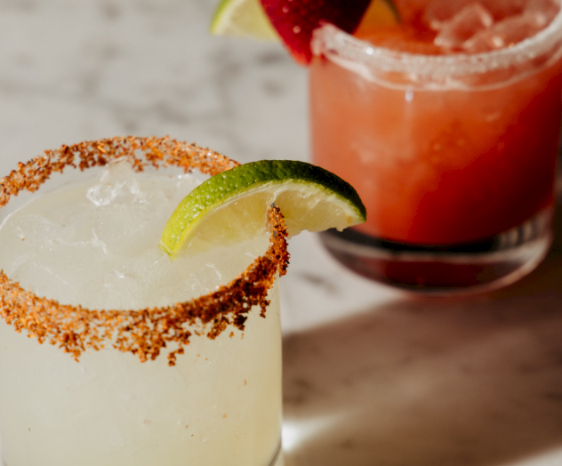 A salt-rimmed cocktail with a lime wedge on the rim, plus a red beverage in a glass in the background, all on a light surface.
