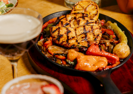 A sizzling skillet with grilled chicken, peppers, and vegetables sits beside cocktails and tacos on a vibrant restaurant table.