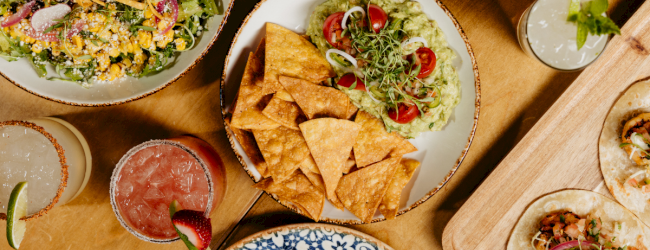 A colorful spread of Mexican-inspired dishes with bowls, chips, salsa, guacamole, and toppings arranged on a wooden table.
