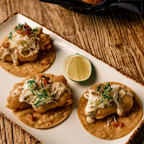 Three tacos on a plate with toppings, lime wedge, and a wooden table background. The scene looks like nachos-style tacos with crema and herbs.