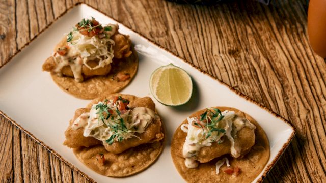 Three tacos on a plate with toppings, lime wedge, and a wooden table background. The scene looks like nachos-style tacos with crema and herbs.