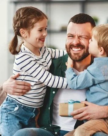 A happy family moment: a bearded dad sits on a couch, warmly hugging his two kids as they smile and share a joyful embrace.