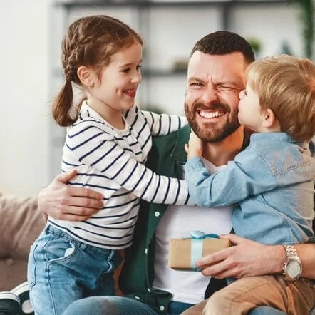 A happy family moment: a bearded dad sits on a couch, warmly hugging his two kids as they smile and share a joyful embrace.