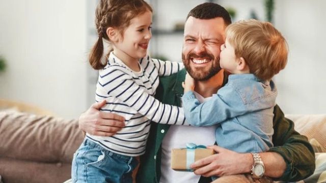 A happy family moment: a bearded dad sits on a couch, warmly hugging his two kids as they smile and share a joyful embrace.