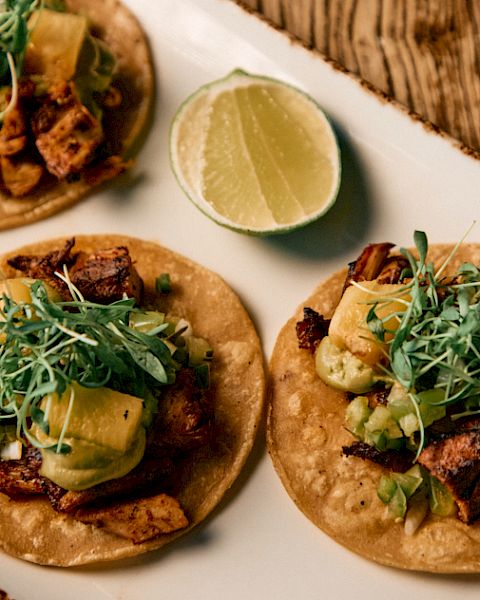 Three tacos on a wooden board, topped with greens, avocado, lime wedge nearby, and crispy meat in wraps.