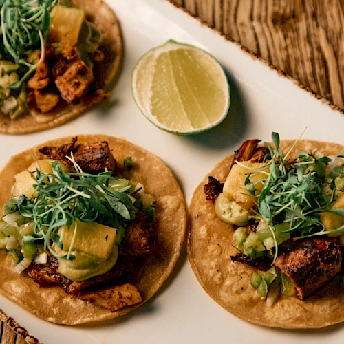 Three tacos on a wooden board, topped with greens, avocado, lime wedge nearby, and crispy meat in wraps.