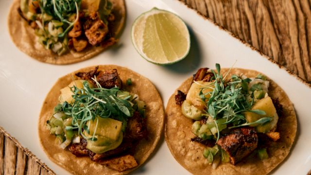 Three tacos on a wooden board, topped with greens, avocado, lime wedge nearby, and crispy meat in wraps.