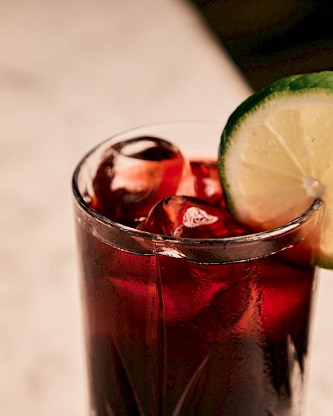 A glass of dark red drink with ice, topped with a lime wheel on the rim, on a light surface.