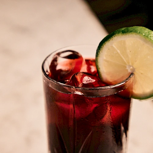 A glass of dark red drink with ice, topped with a lime wheel on the rim, on a light surface.