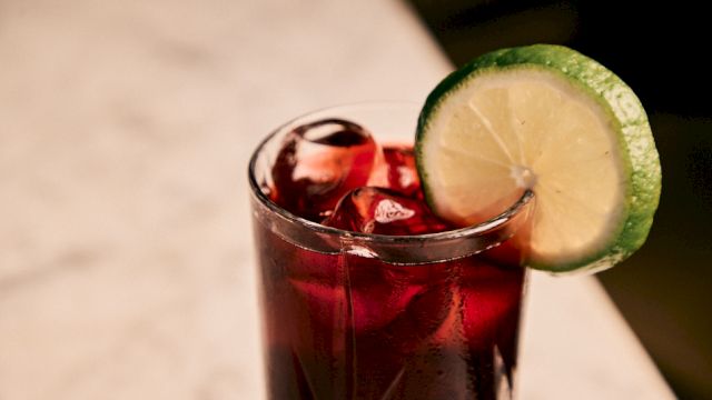 A glass of dark red drink with ice, topped with a lime wheel on the rim, on a light surface.