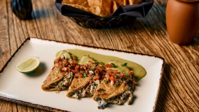 Crispy fish tacos with salsa, lime wedge, roasted corn tortilla chips, and a mug of coffee on a rustic wooden table.