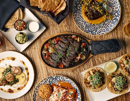 A spread of Mexican-inspired dishes: grilled meats, tacos with toppings, a sizzling skillet, bowls with sauces, tortillas, and fresh garnishes on a rustic wooden table.