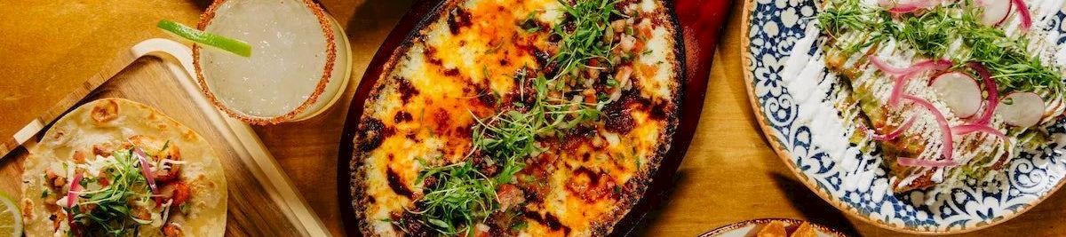A table with a variety of dishes: a skillet of cheesy baked dish, salads, tacos/mini wraps, tortilla chips with salsa, and a fresh herb garnish, all on wooden and ceramic plates.