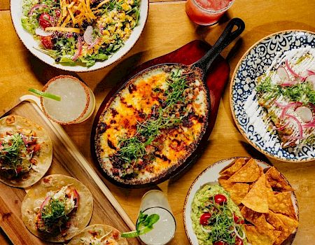 A table with a variety of dishes: a skillet of cheesy baked dish, salads, tacos/mini wraps, tortilla chips with salsa, and a fresh herb garnish, all on wooden and ceramic plates.