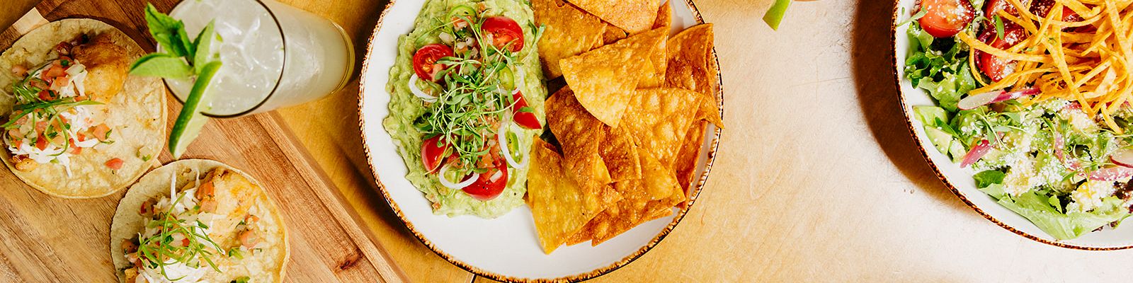 A colorful spread of Mexican-inspired foods: bowls of nachos, guacamole, tacos, tortilla chips, a salad, and drinks on a wooden table.