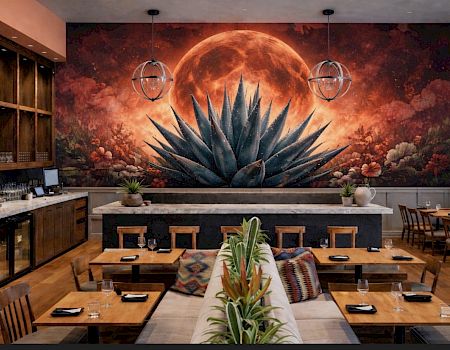 A stylish dining area with warm wood tables, a long central plant arrangement, and a dramatic wall mural of a glowing red moon over an agave plant, flanked by globe pendant lights. Top it at 140 characters, always ending the sentence.
