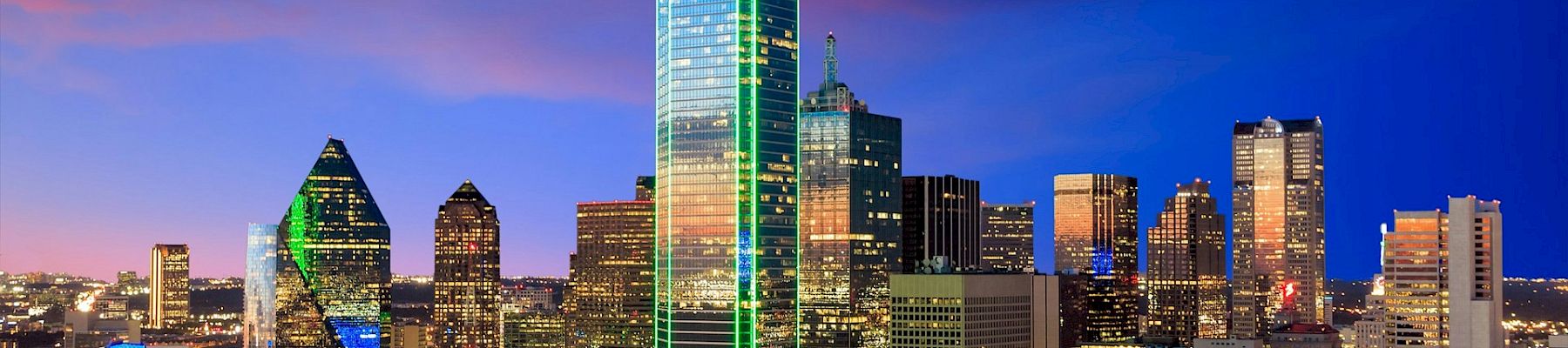 A nighttime city skyline with tall illuminated skyscrapers glowing against a deep blue sky, reflecting urban lights and modern architecture.