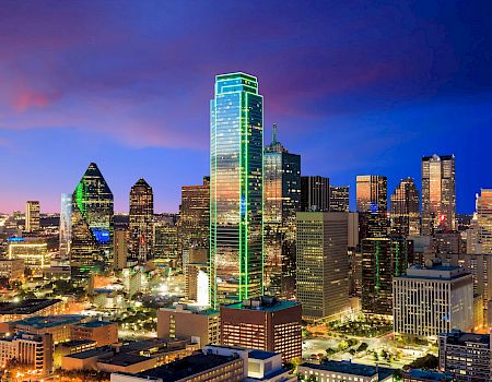 A nighttime city skyline with tall illuminated skyscrapers glowing against a deep blue sky, reflecting urban lights and modern architecture.
