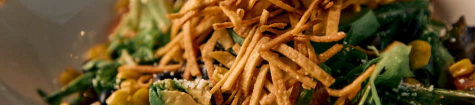 A colorful salad with greens, cherry tomato halves, corn, avocado, and herbs, topped with crisp fried sticks and mixed with crunchy twists.