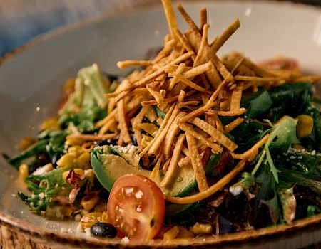 A colorful salad with greens, cherry tomato halves, corn, avocado, and herbs, topped with crisp fried sticks and mixed with crunchy twists.