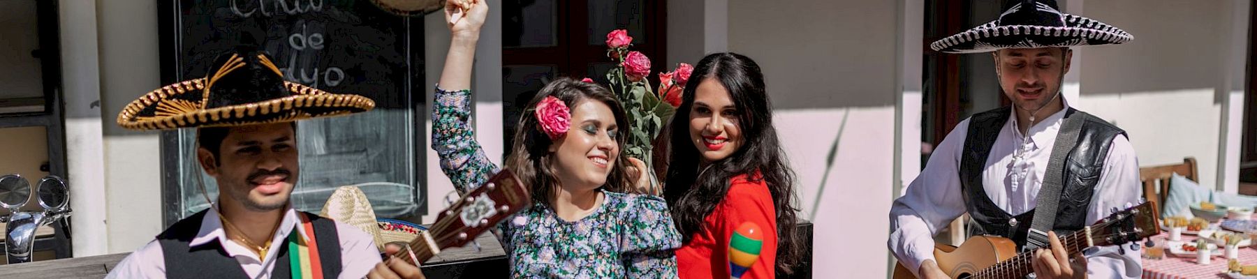 A lively trio and two guitarists in a festive outdoor setting, two women in colorful dresses clap and pose with maracas, sombreros, and guitars.