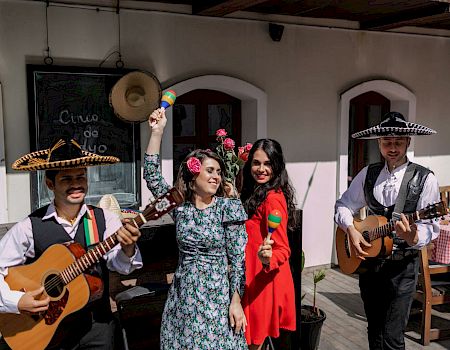 A lively trio and two guitarists in a festive outdoor setting, two women in colorful dresses clap and pose with maracas, sombreros, and guitars.