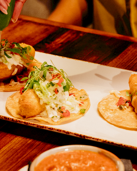 Mini tacos with fried shells topped with shredded cabbage, pico de gallo, and greens, with sauce being poured on.