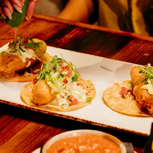 Mini tacos with fried shells topped with shredded cabbage, pico de gallo, and greens, with sauce being poured on.