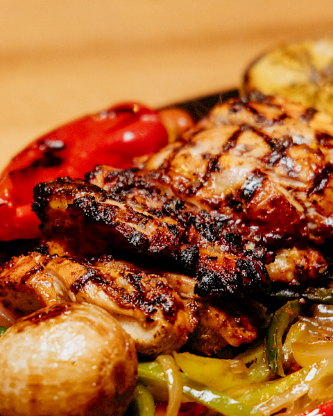 A sizzling grilled meat topped with char and herbs, served in a skillet with colorful roasted peppers, onions, and potatoes.