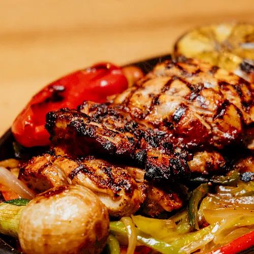 A sizzling grilled meat topped with char and herbs, served in a skillet with colorful roasted peppers, onions, and potatoes.