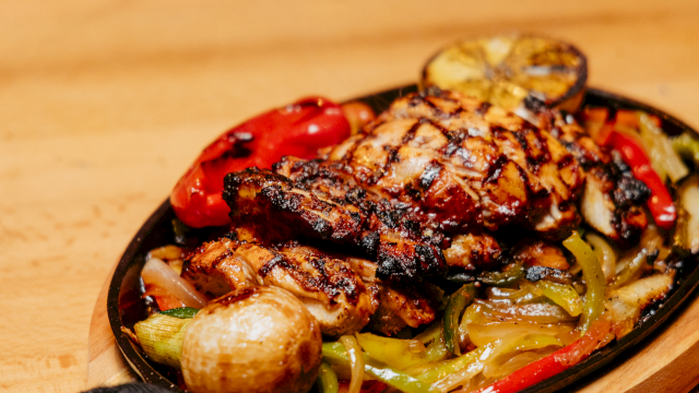 A sizzling grilled meat topped with char and herbs, served in a skillet with colorful roasted peppers, onions, and potatoes.