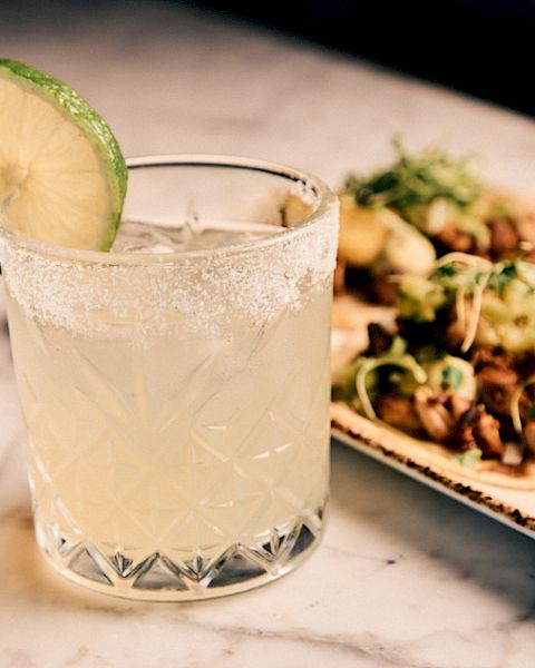 A frosty cocktail in a crystal glass with a lime slice, beside a platter of tacos with lime wedges and herbs.