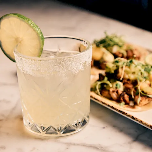 A frosty cocktail in a crystal glass with a lime slice, beside a platter of tacos with lime wedges and herbs.