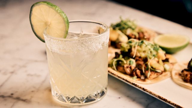 A frosty cocktail in a crystal glass with a lime slice, beside a platter of tacos with lime wedges and herbs.