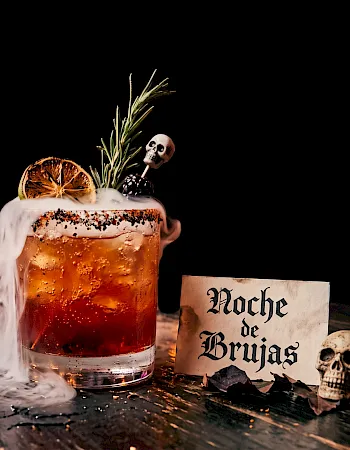 A spooky Halloween beer: a frothy mug with smoke, rosemary, lemon, skull decor, and a sign reading &ldquo;Noche de Brujas.&rdquo;