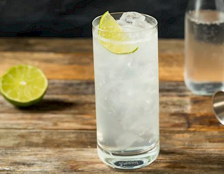 A clear lime-and-ice cocktail in a tall glass sits on a wooden bar with lime wedges, plus a jigger and shot glass.