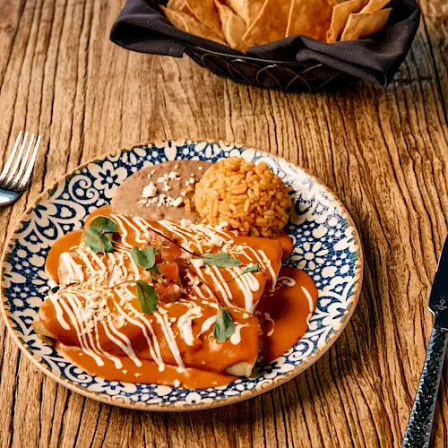 A plate of orange-sauced dumplings with rice and basil sits on a wooden table beside forks, knife, and a napkin.