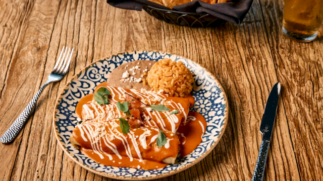 A plate of orange-sauced dumplings with rice and basil sits on a wooden table beside forks, knife, and a napkin.